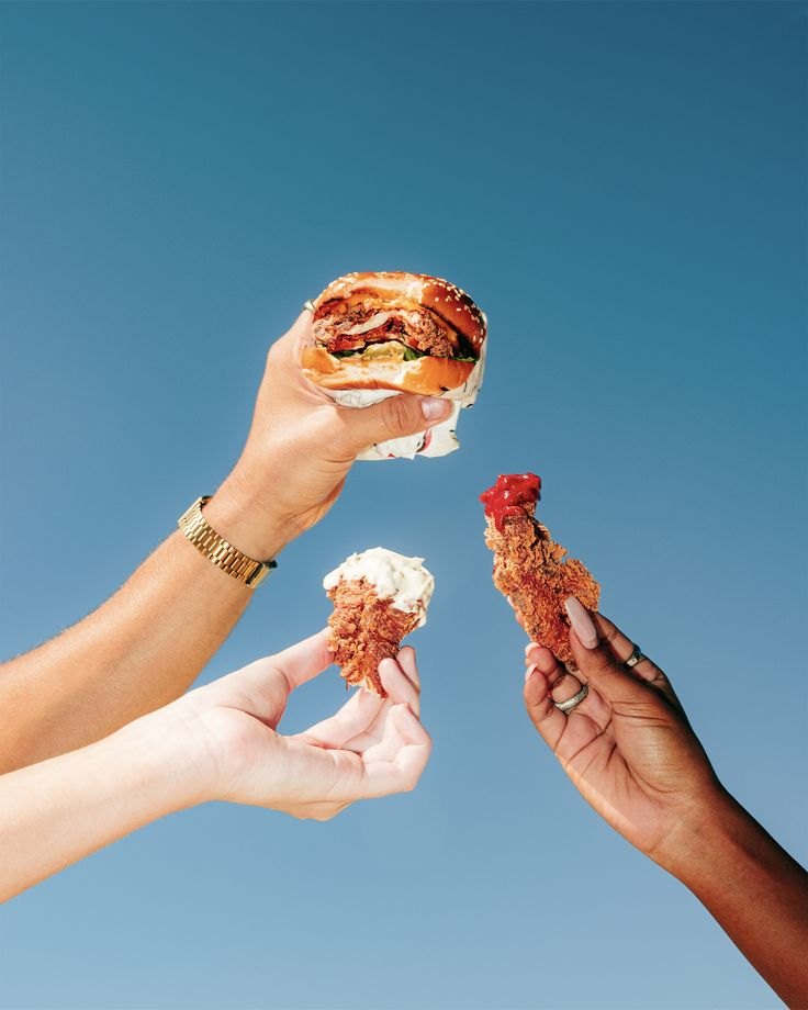 Three hands holding a burger and fried chicken against the sky